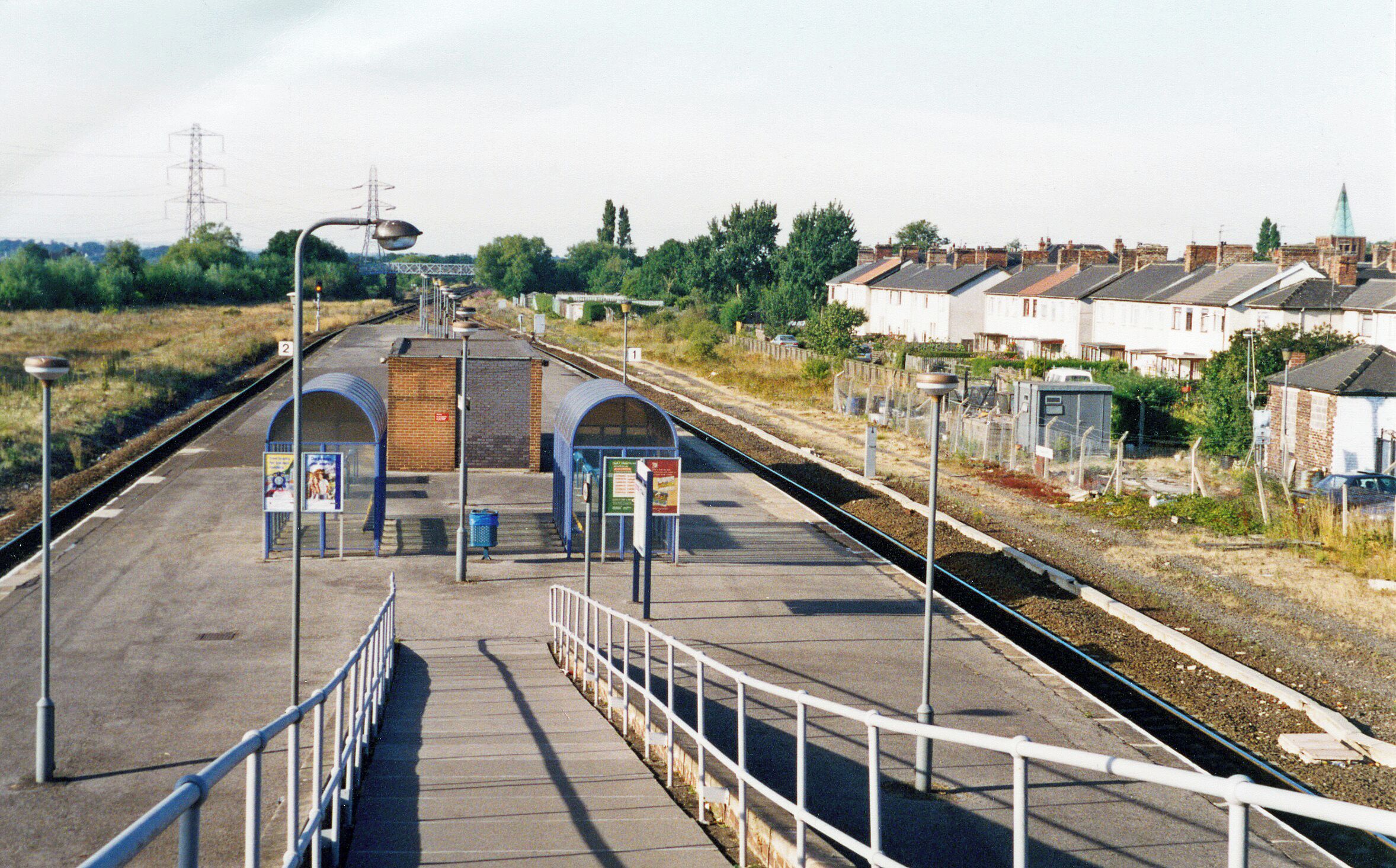 A railway station platform with a pedestrian bridge, a signal tower, and residential houses in the background.
