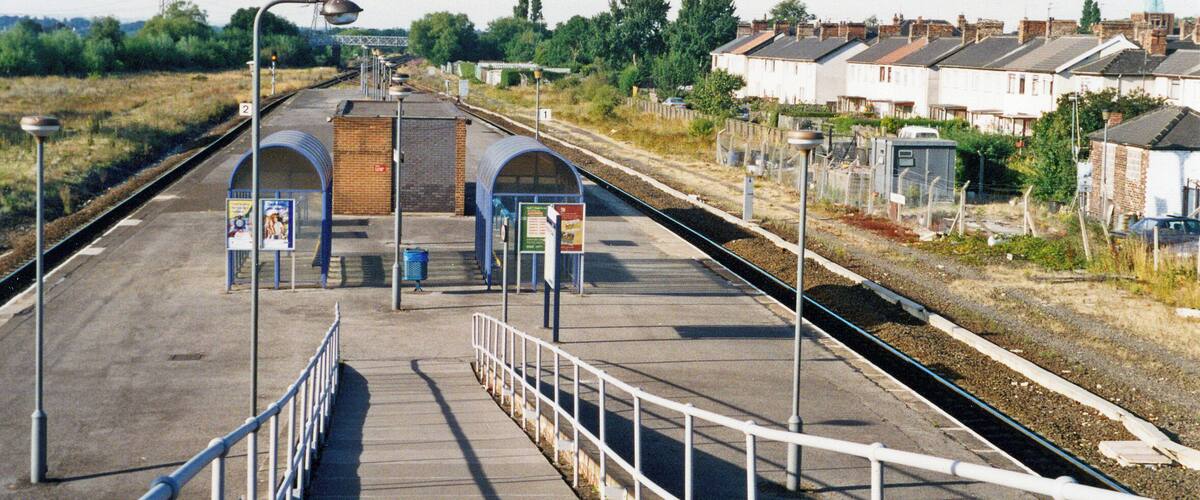 A railway station platform with a pedestrian bridge, a signal tower, and residential houses in the background.