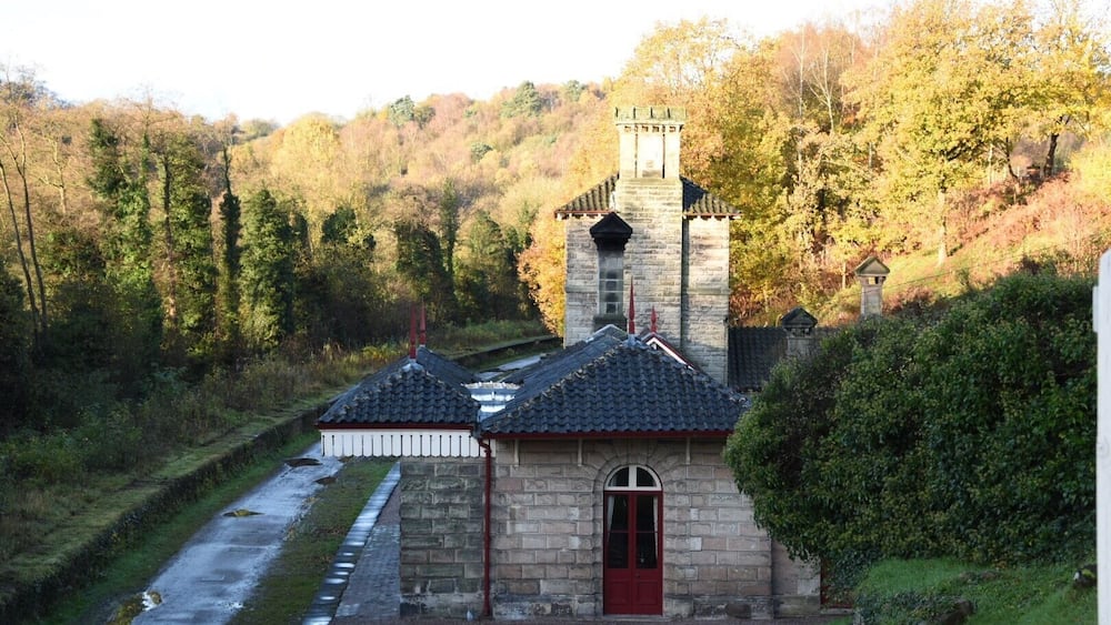 Un bâtiment en pierre avec une porte rouge et une tour, entouré d’arbres et bordé par une rivière.