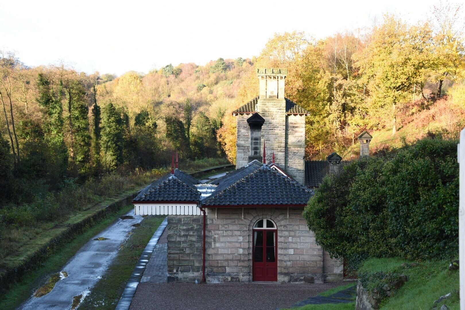 A stone building with a red door and a tower, surrounded by trees and a river.