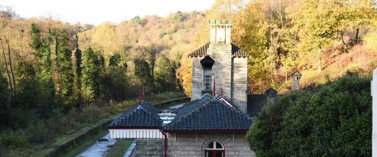 A stone building with a red door and a tower, surrounded by trees and a river.