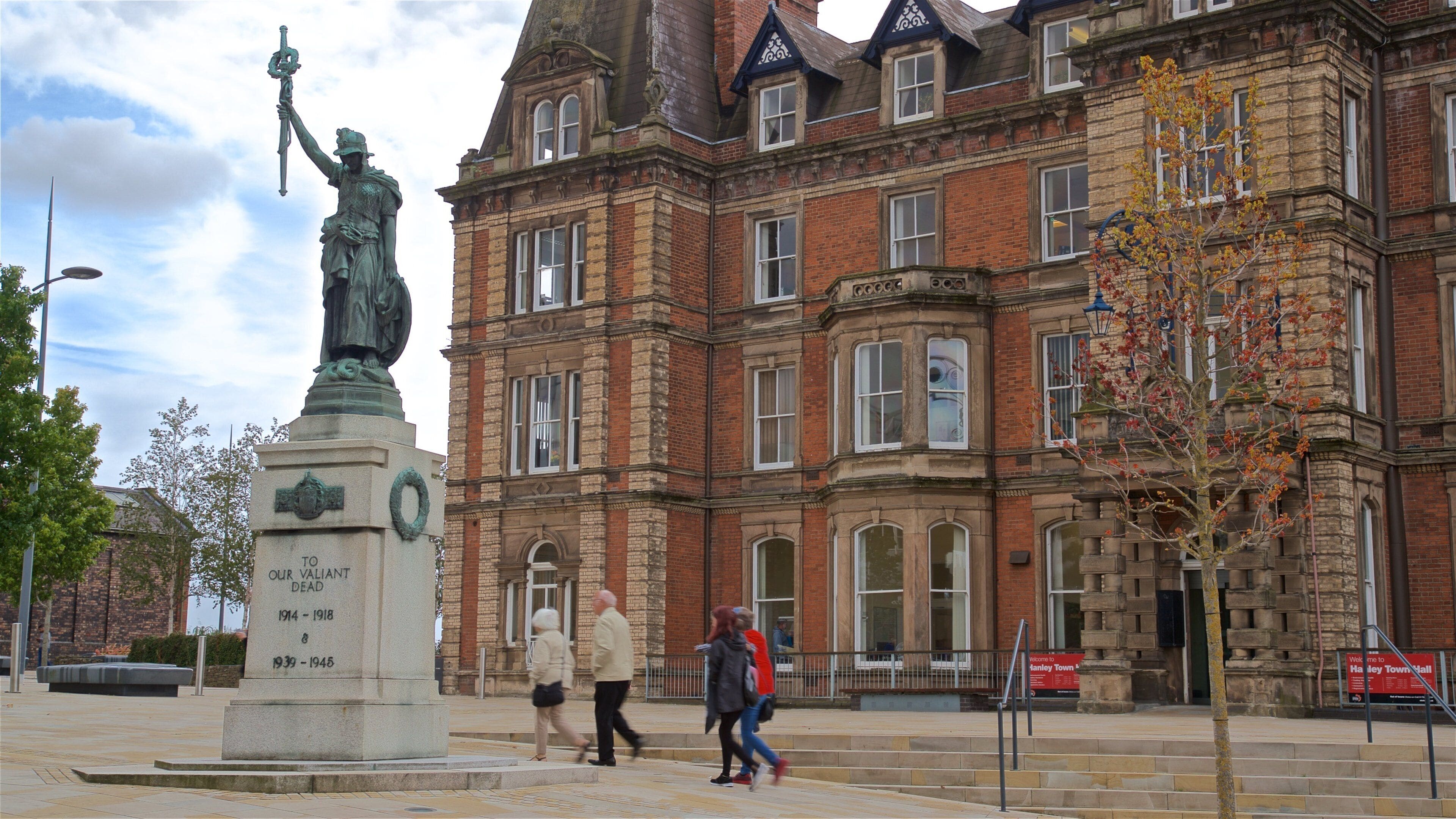 A statue of a figure holding a sword, a red brick building with multiple windows, and a sign that reads "Welcome to Hanley Town Hall".