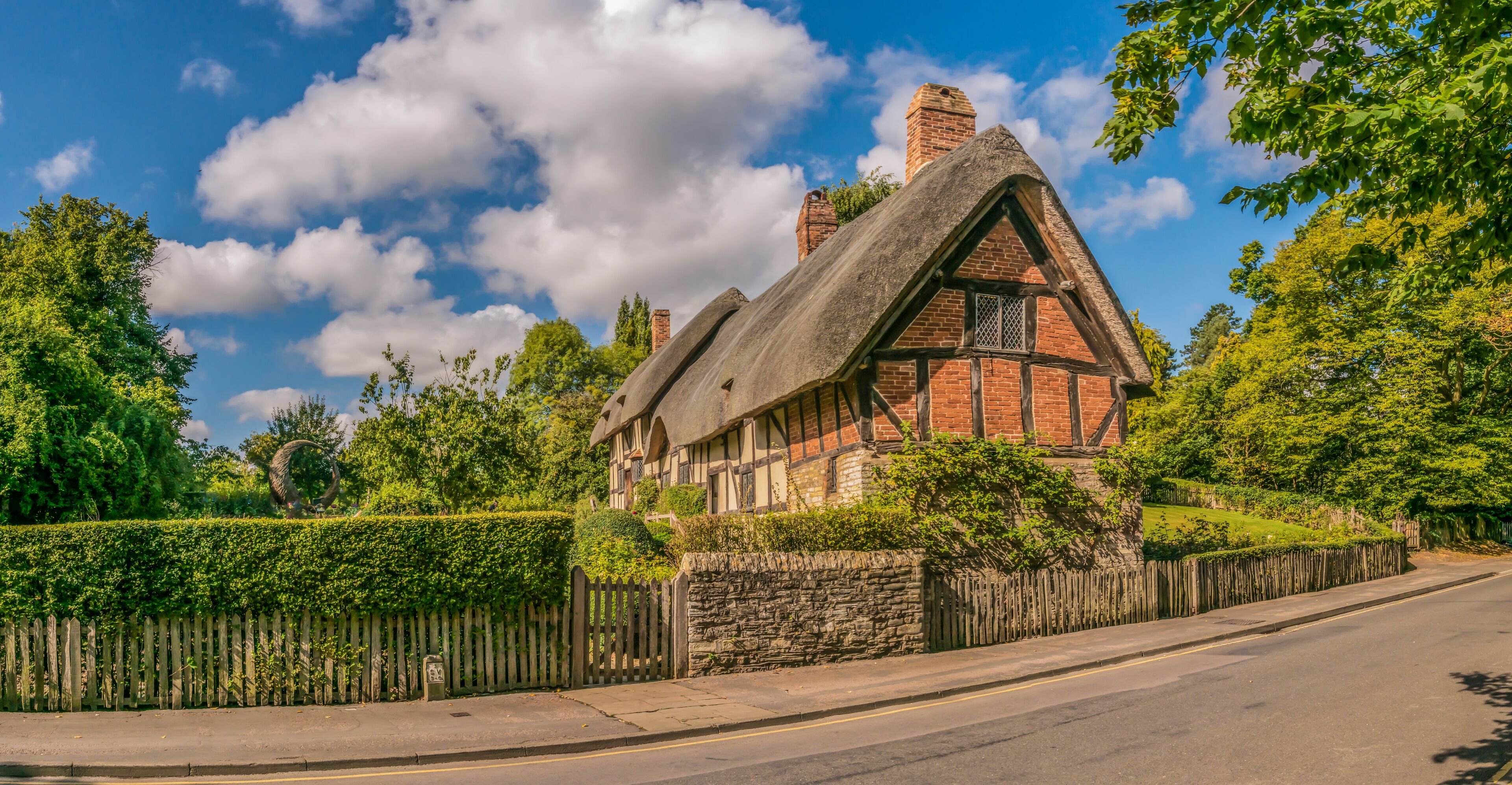 A traditional half-timbered house with a steeply pitched roof, surrounded by lush greenery and a wooden fence.