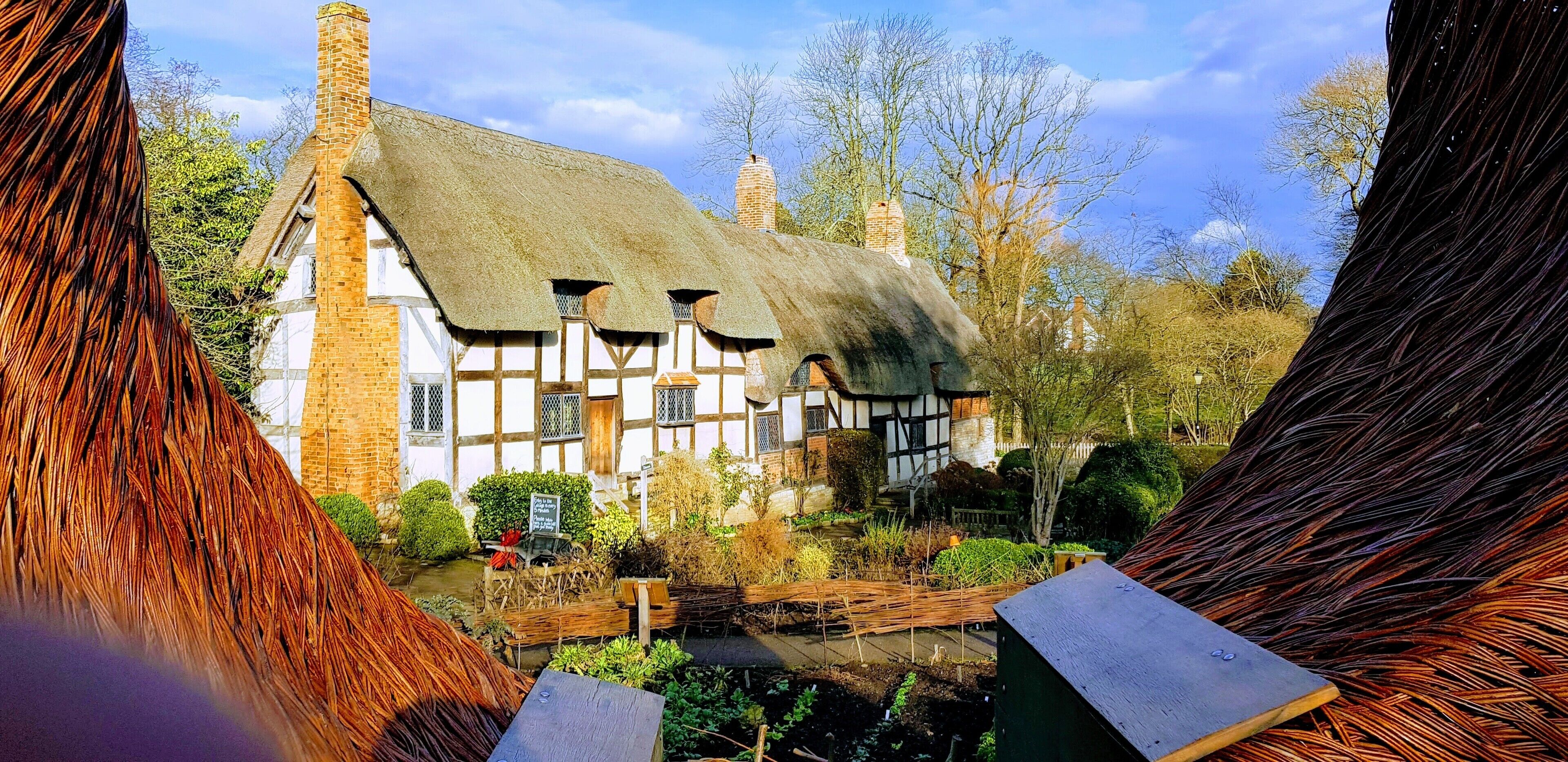 A traditional half-timbered house with a thatched roof, surrounded by a garden and trees.
