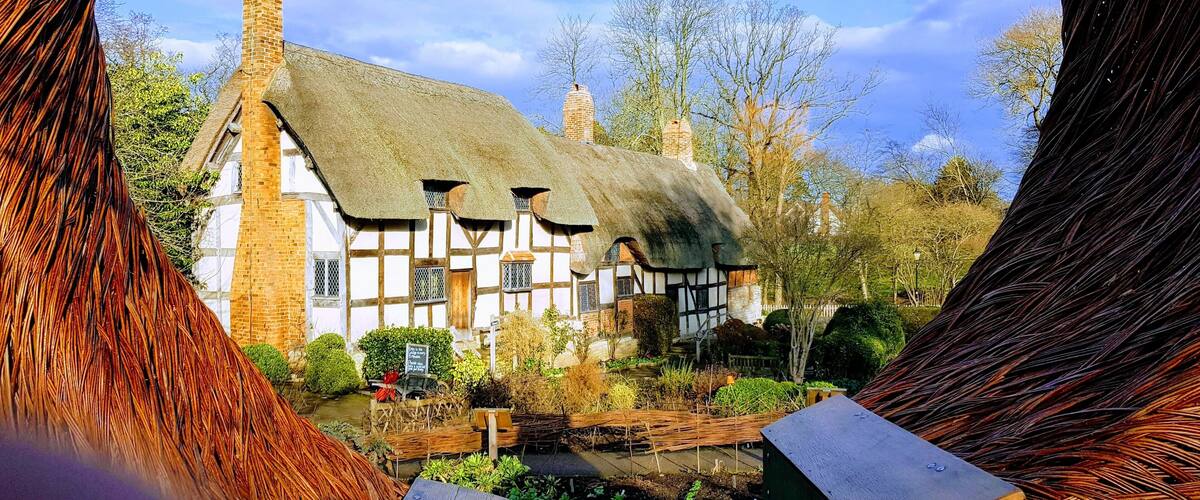 A traditional half-timbered house with a thatched roof, surrounded by a garden and trees.