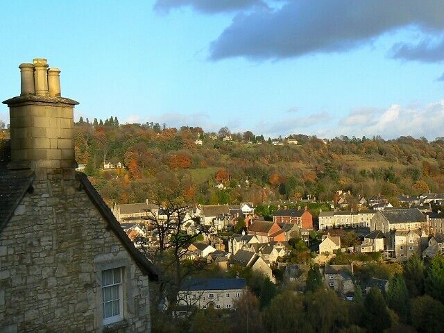 A town with stone buildings, a hill with autumn-colored trees, and a clear sky.