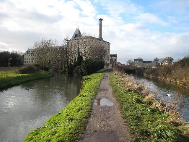A canal with a path alongside, a historic building with a tower, and a bridge in the background.