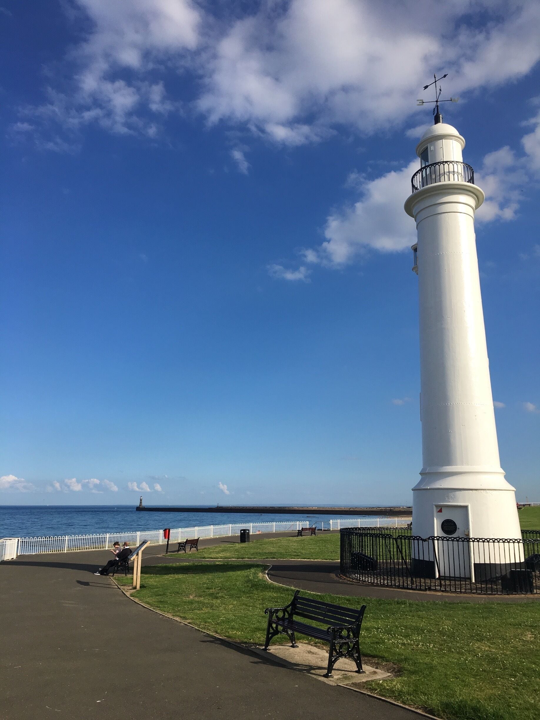 A white lighthouse with a weather vane on top, a bench, and a white fence along the shoreline.