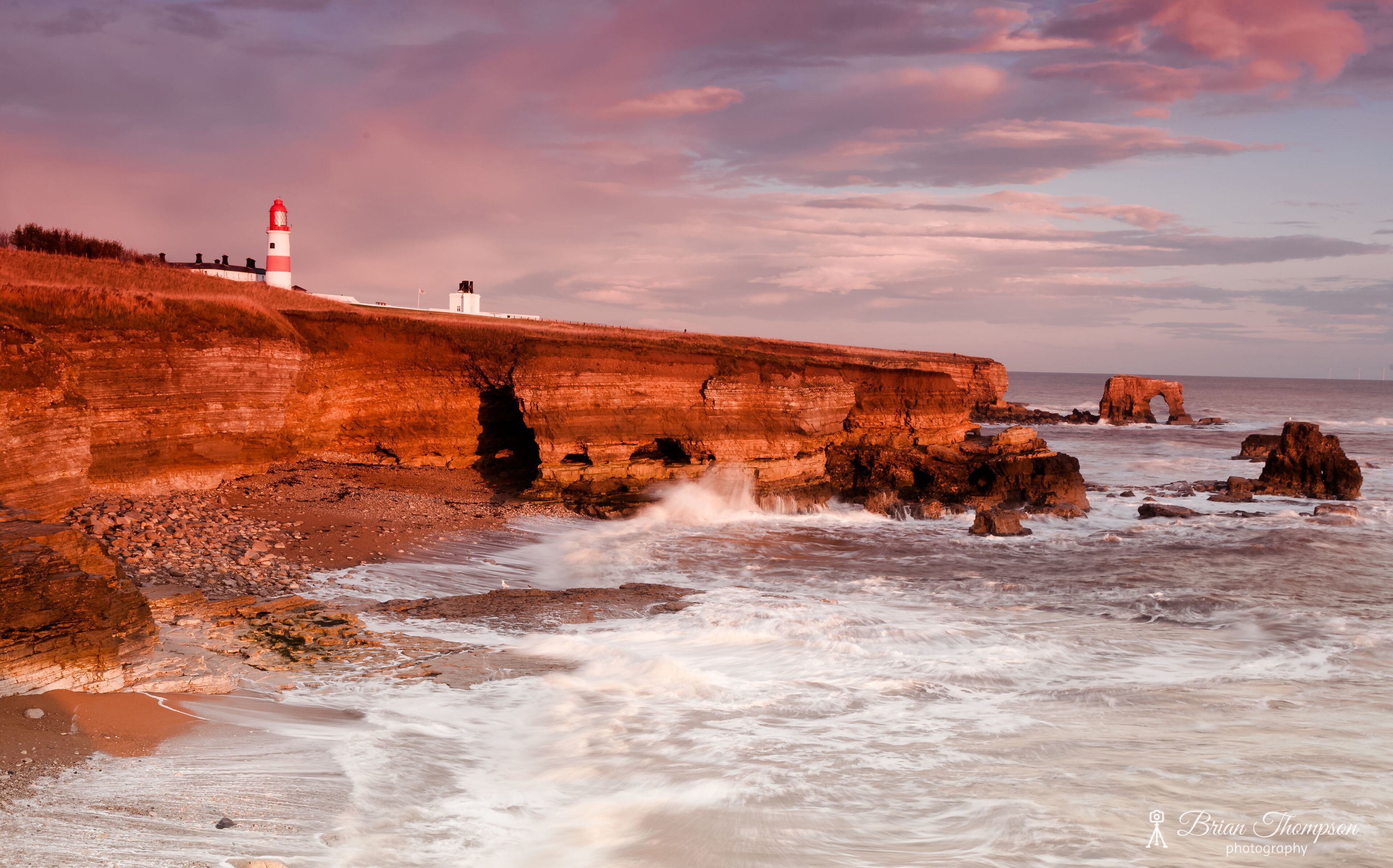 A coastal landscape with a lighthouse, rocky cliffs, and waves crashing against the shore.