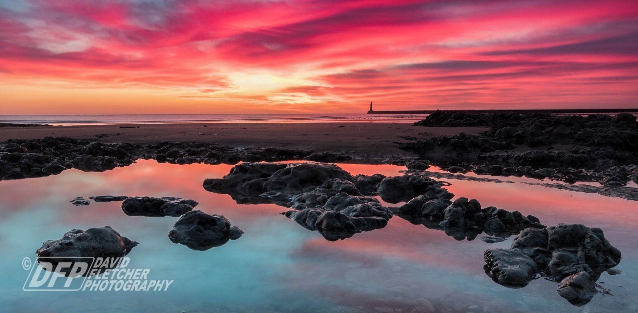 A rocky shoreline with a lighthouse in the distance during sunset.
