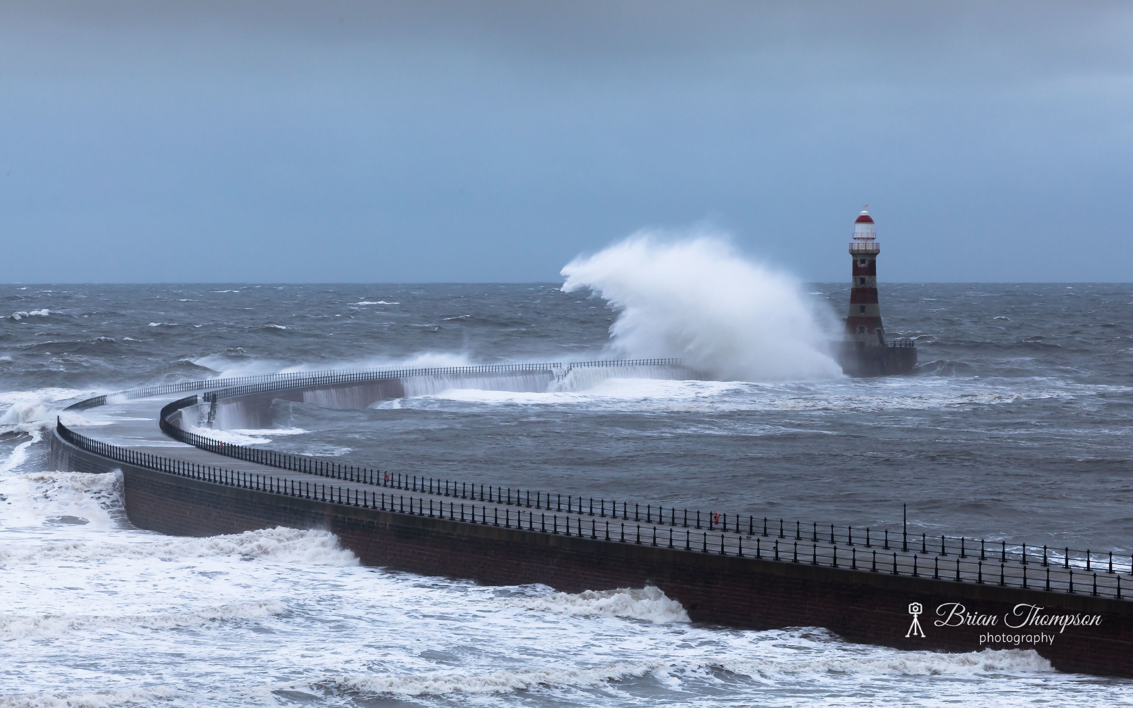 A lighthouse and a sea wall with waves crashing against it.