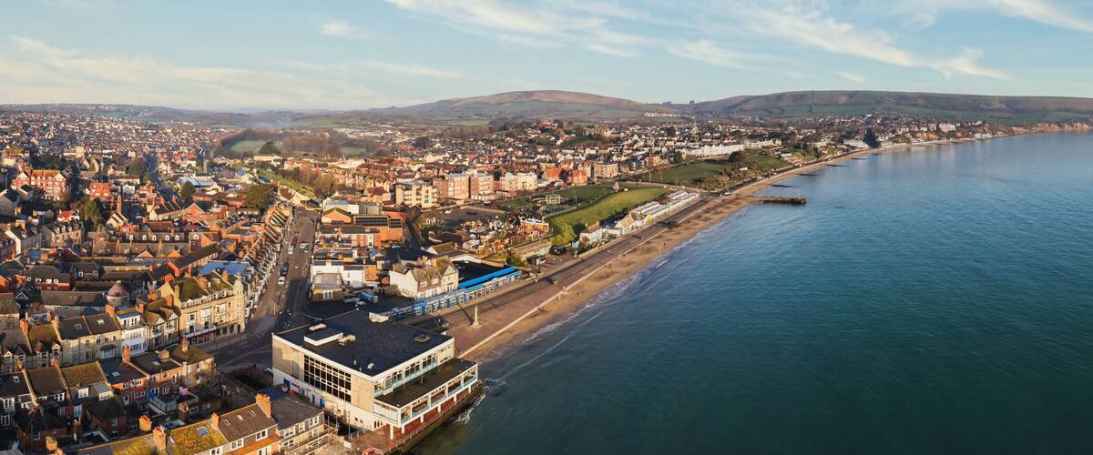A coastal town with a waterfront promenade, a large building with a blue roof, and rolling hills in the background.