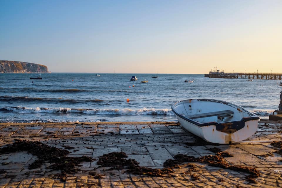 A coastal scene with a boat on a paved jetty, calm sea, distant cliffs, and a pier extending into the water.