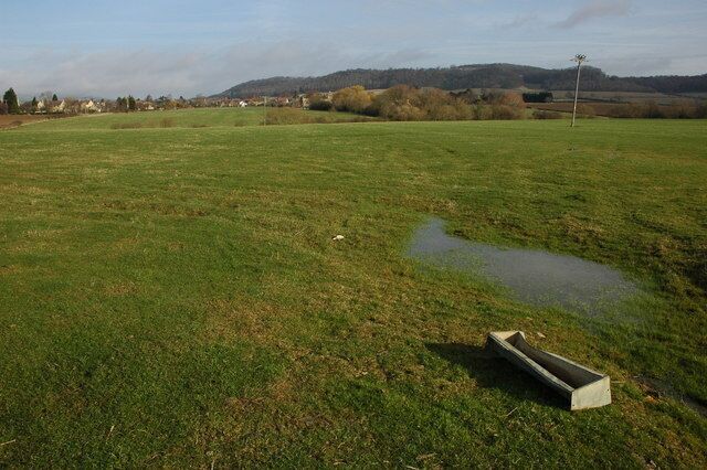 Fields at Alderton Grazing land to the south of Alderton, the village and the church can be seen in the middle distance. Alderton Hill, a Cotswold outlier can be seen in the background. The far side the hill is called Dumbleton Hill, it is also less widely known as Oak Hill. In the foreground is a sheep feeding trough.