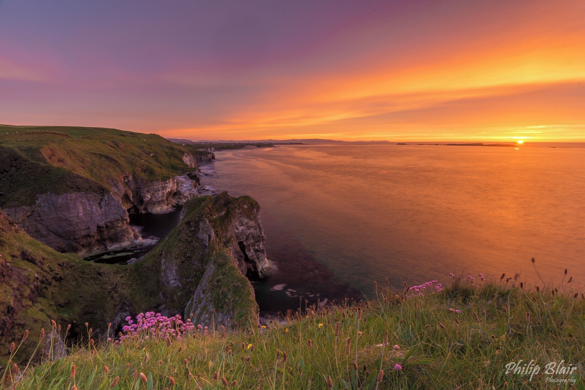A coastal landscape with cliffs, grassy areas, and a sunset over the water.