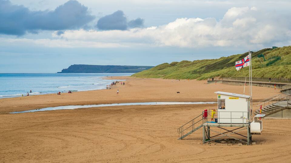A sandy beach with a lifeguard tower, grassy dunes, and a distant cliff.