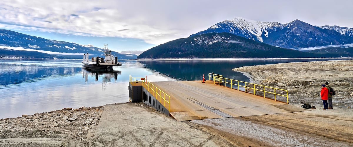 A calm lake with a dock, a ship, and snow-capped mountains in the background.