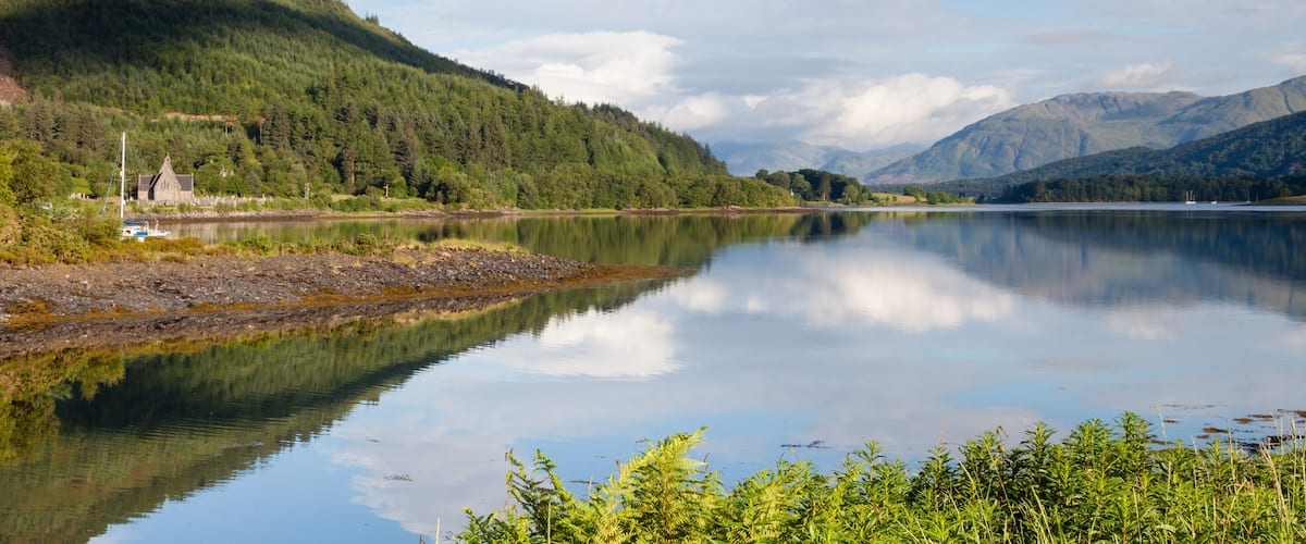 A calm lake with a forested mountain in the background and a small building near the shore.