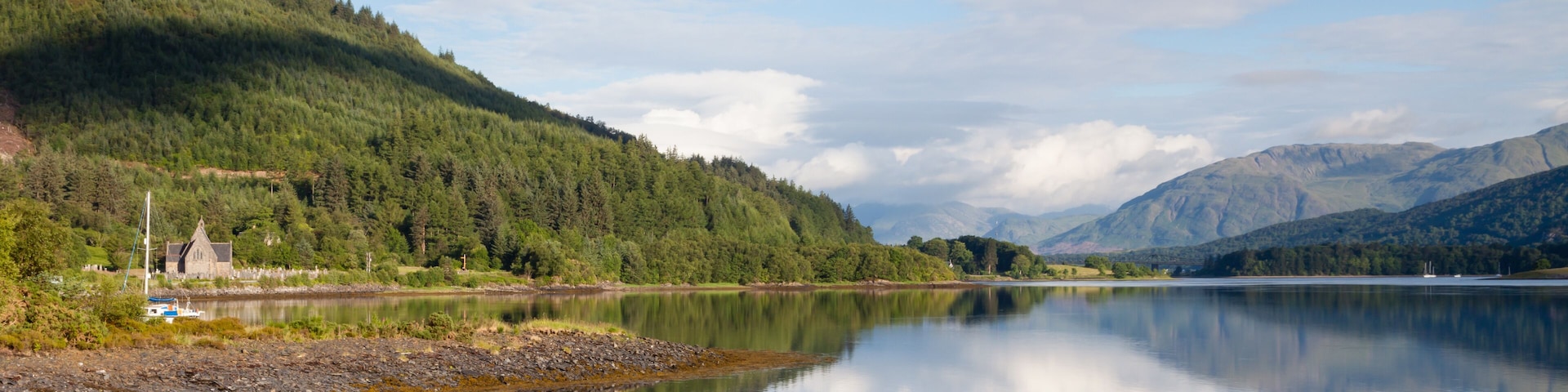 A calm lake with a forested mountain in the background and a small building near the shore.
