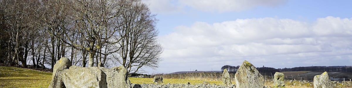 Ein Steinkreis mit hoch aufragenden Steinen in einem grasbewachsen Feld, umgeben von baumlosen Bäumen und einem teils bewölkten Himmel.