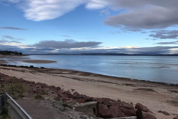 Paisagem costeira com uma praia de areia, afloramentos rochosos e um mar calmo sob um céu parcialmente nublado.