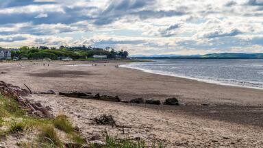 A beach with a rocky shoreline, a distant shoreline with buildings, and a cloudy sky.