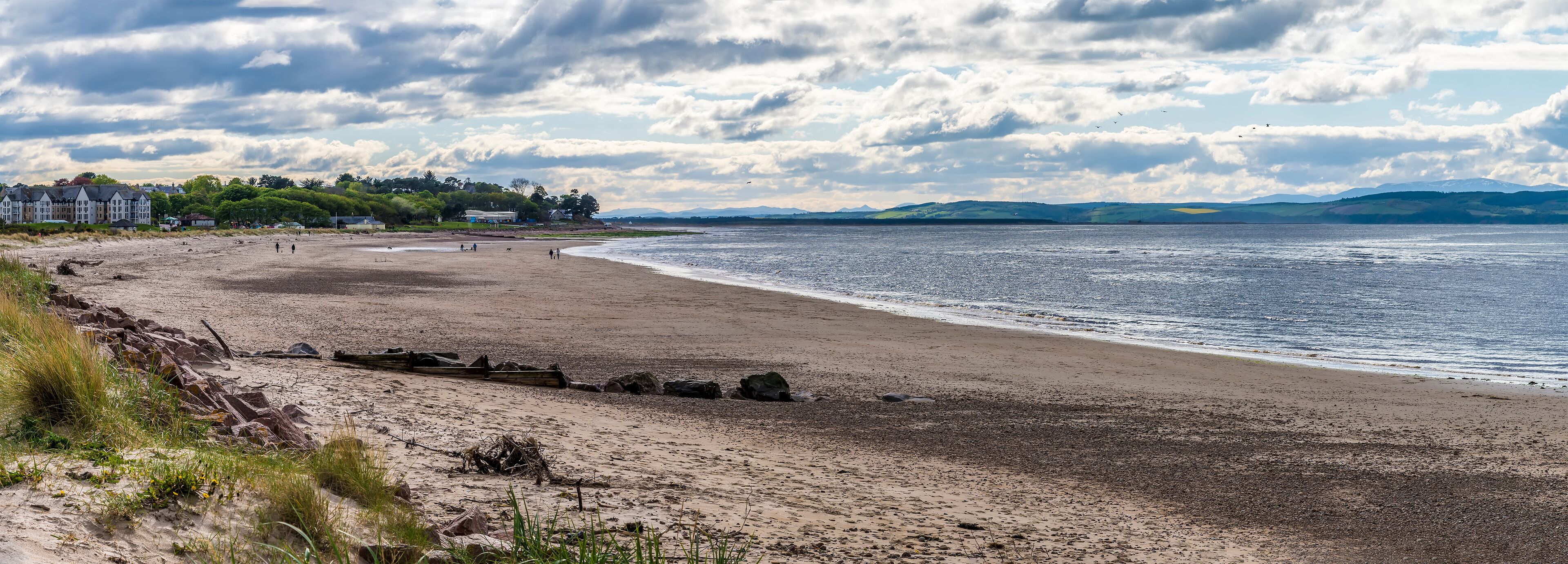 A beach with a rocky shoreline, a distant shoreline with buildings, and a cloudy sky.