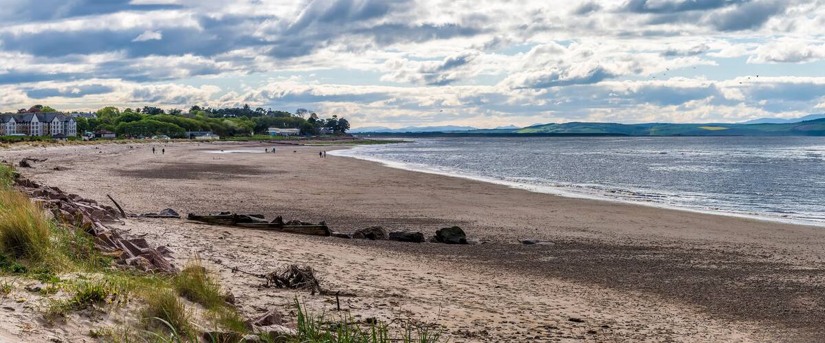 A beach with a rocky shoreline, a distant shoreline with buildings, and a cloudy sky.