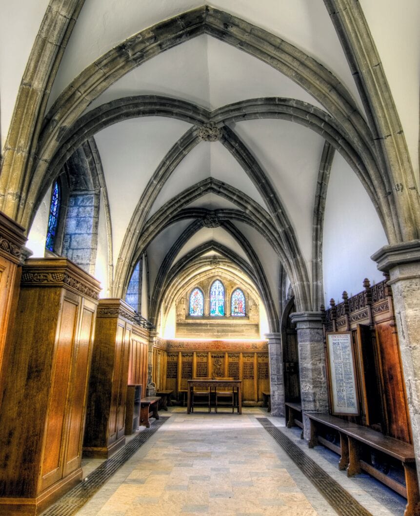A church interior with high vaulted ceilings, wooden pews, and stained glass windows.