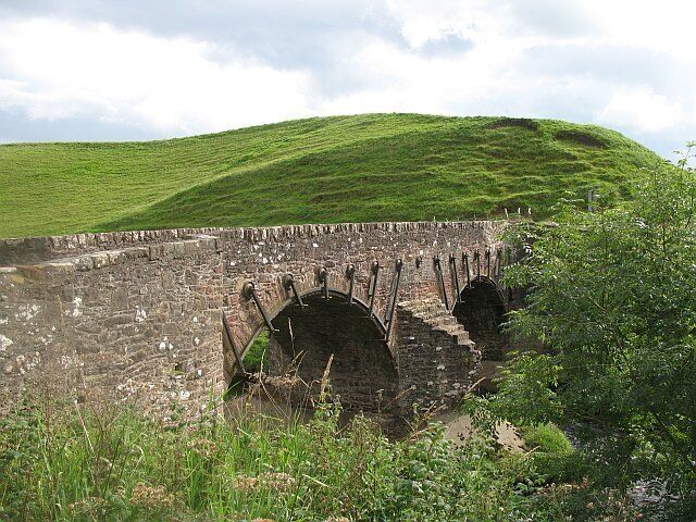 A stone bridge over a water channel, surrounded by lush green hills.