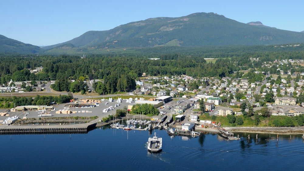 A coastal town with a marina, surrounded by forested hills and mountains.