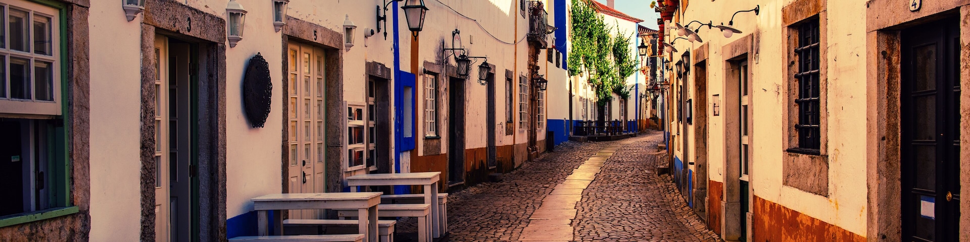 Una calle adoquinada flanqueada por edificios blancos, algunos con detalles azules, y zonas con mesas al aire libre.