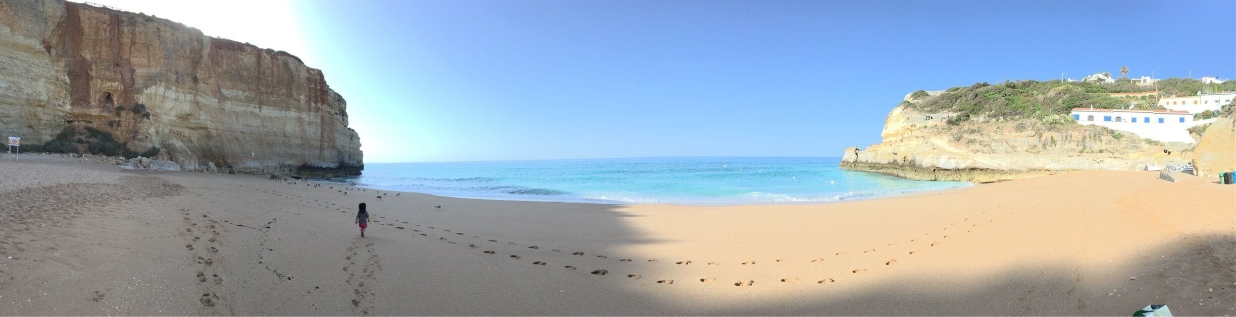 A beach with footprints leading to the water, a cliff, and buildings on a hillside.
