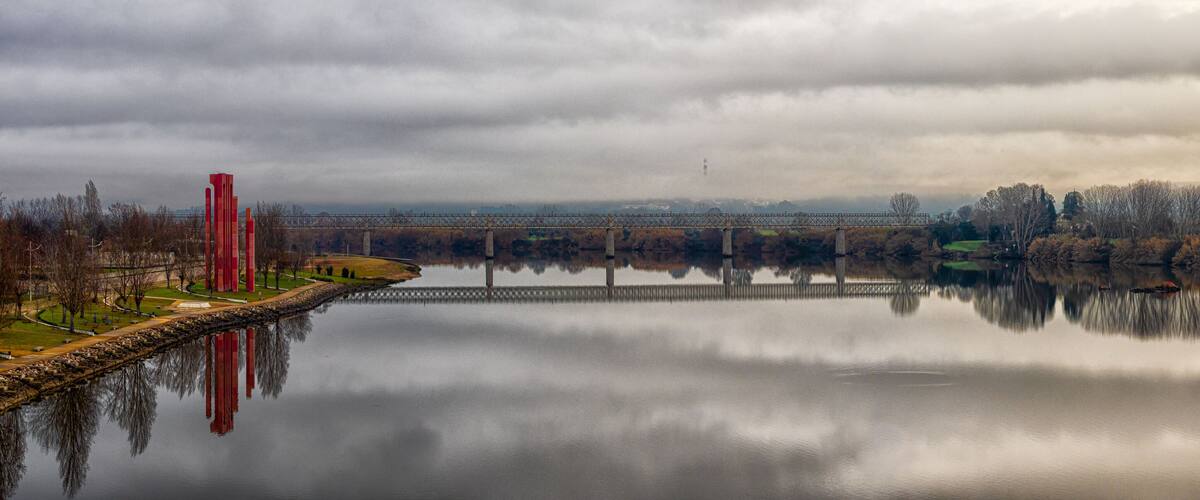 A calm river with a bridge, red sculptures, and a cloudy sky.