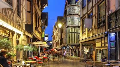Una calle estrecha flanqueada por edificios tradicionales con entramado de madera, una torre destacada con techo cónico y zonas de estar al aire libre con mesas y sillas.