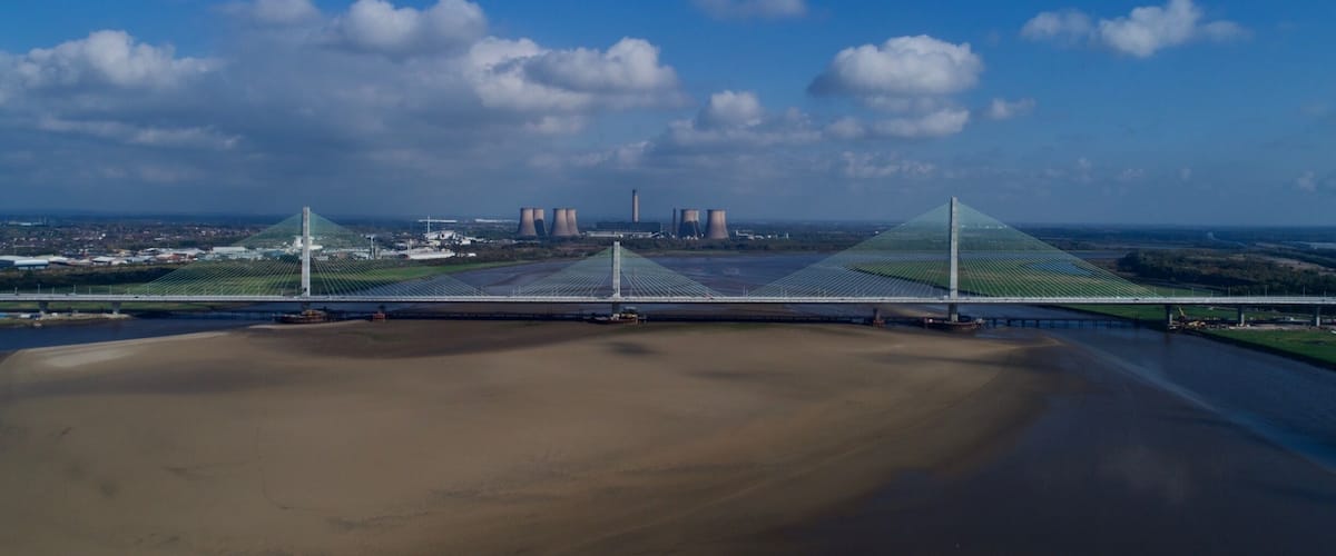 A bridge with a unique cable design, a river in the foreground, and cooling towers in the background.