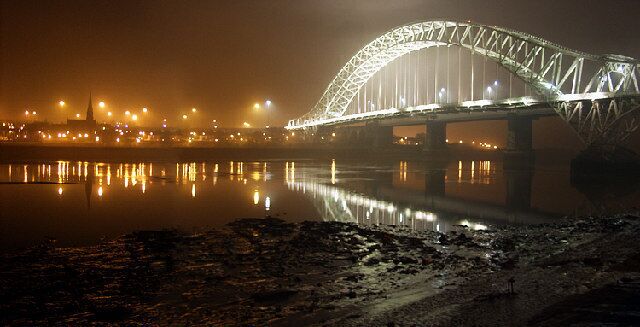 A nighttime view of a large arched bridge over a river, with city lights reflecting on the water.