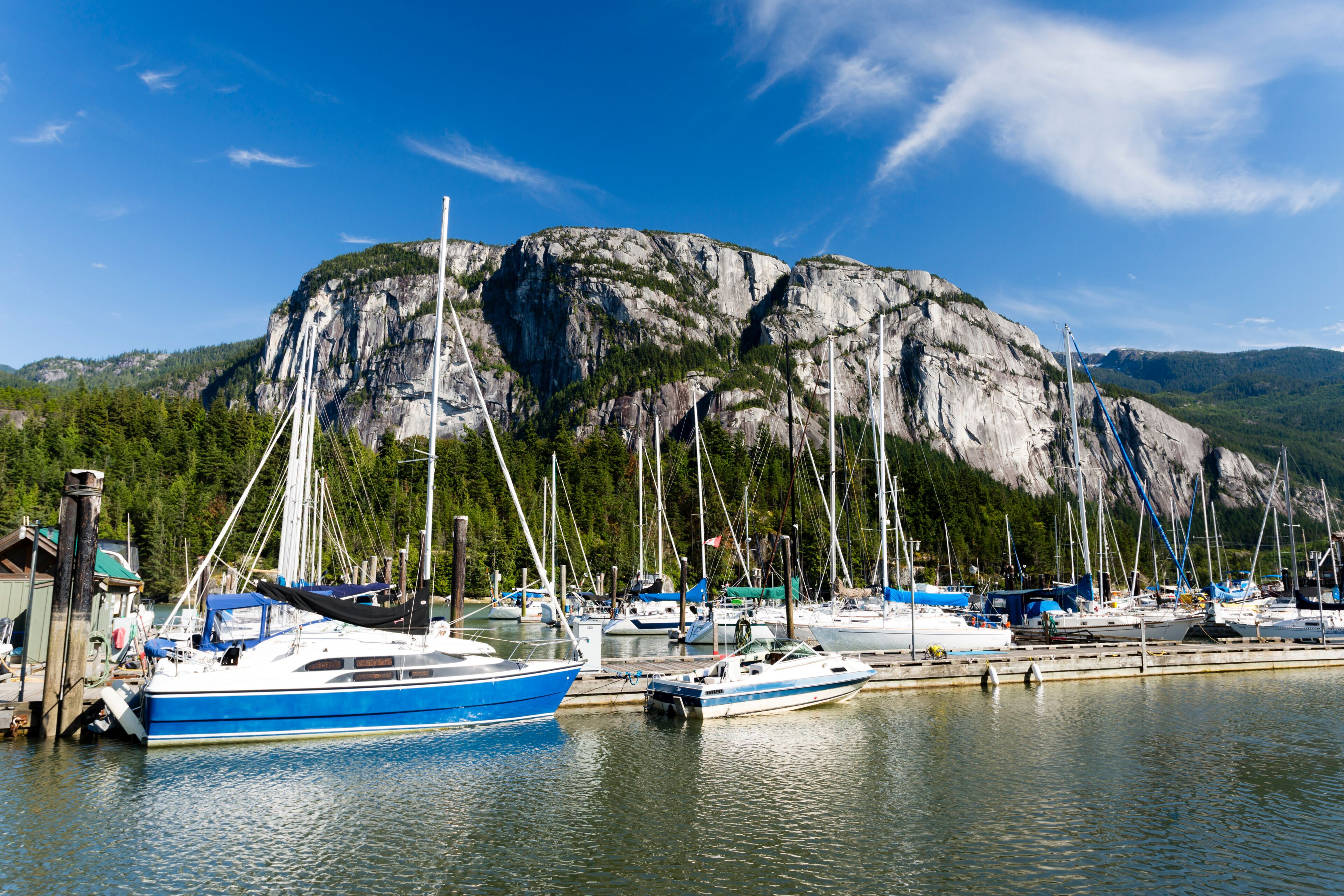 Marina con barcos amarrados en aguas tranquilas, un acantilado rocoso grande al fondo y un cielo azul despejado.