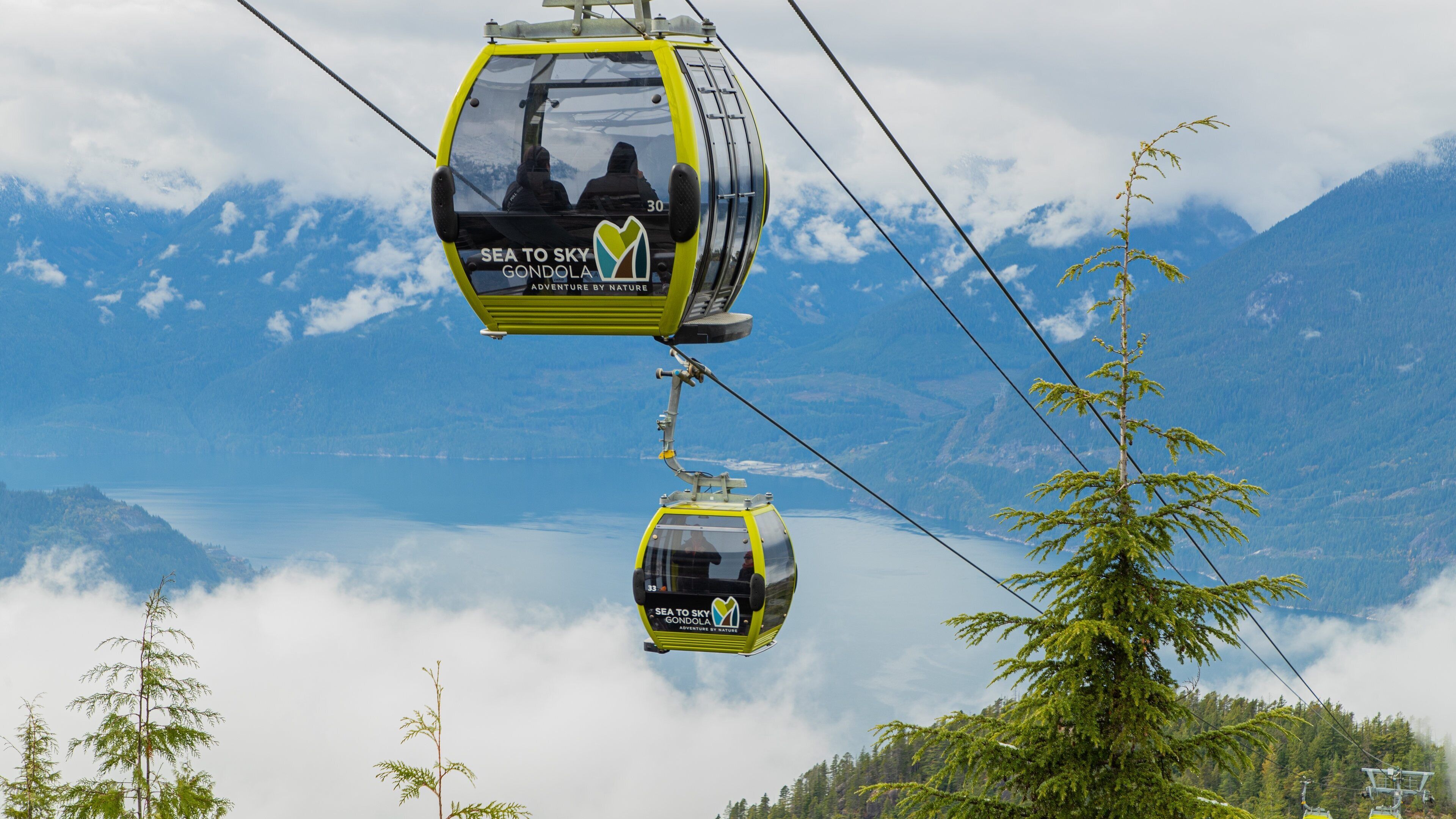 A cable car with "Sea to Sky Gondola" branding, flying over a mountainous landscape with a lake and forested areas.