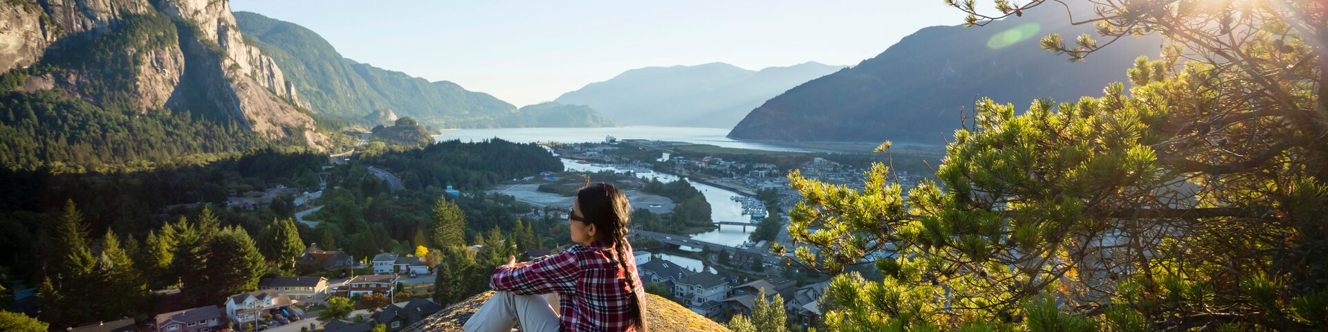 Una persona sentada sobre un saliente rocoso contemplando un valle con un río y montañas al fondo.