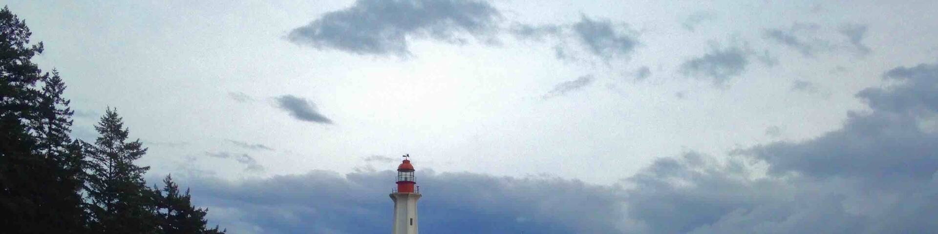 Un phare se dresse sur une île rocheuse, coiffé d’un toit rouge, et est entouré d’eaux calmes et d’une forêt dense.