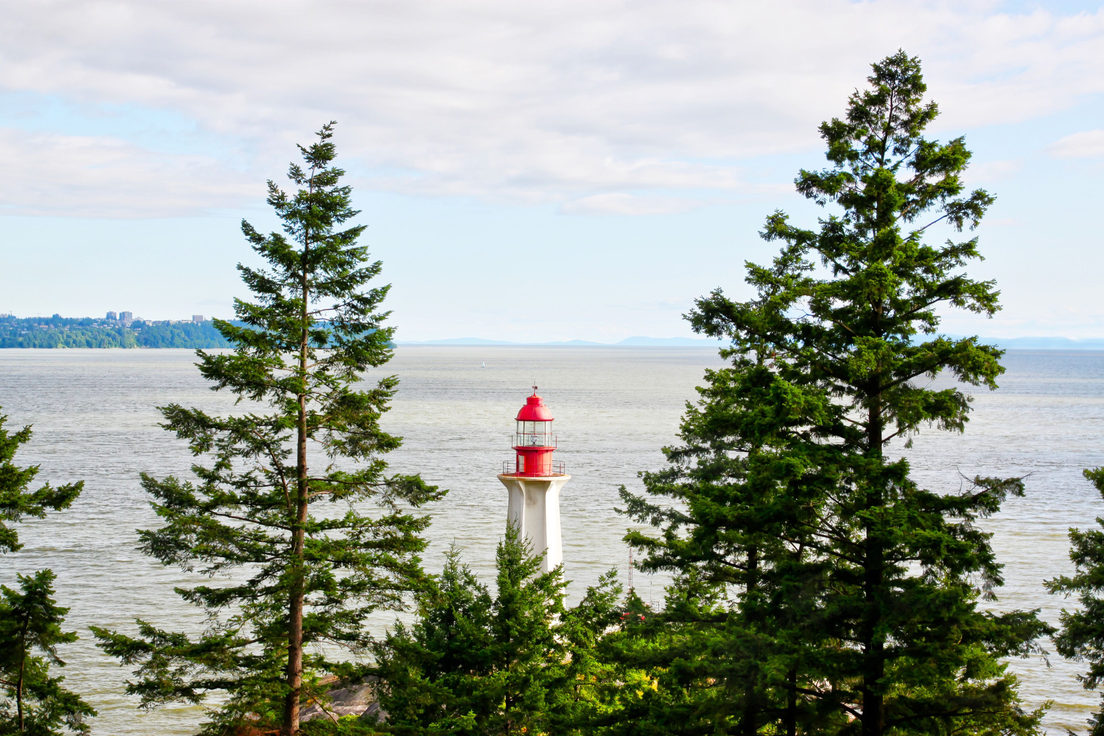 Un faro rodeado de árboles de hoja perenne, con vistas a una masa de agua tranquila y un horizonte urbano distante.