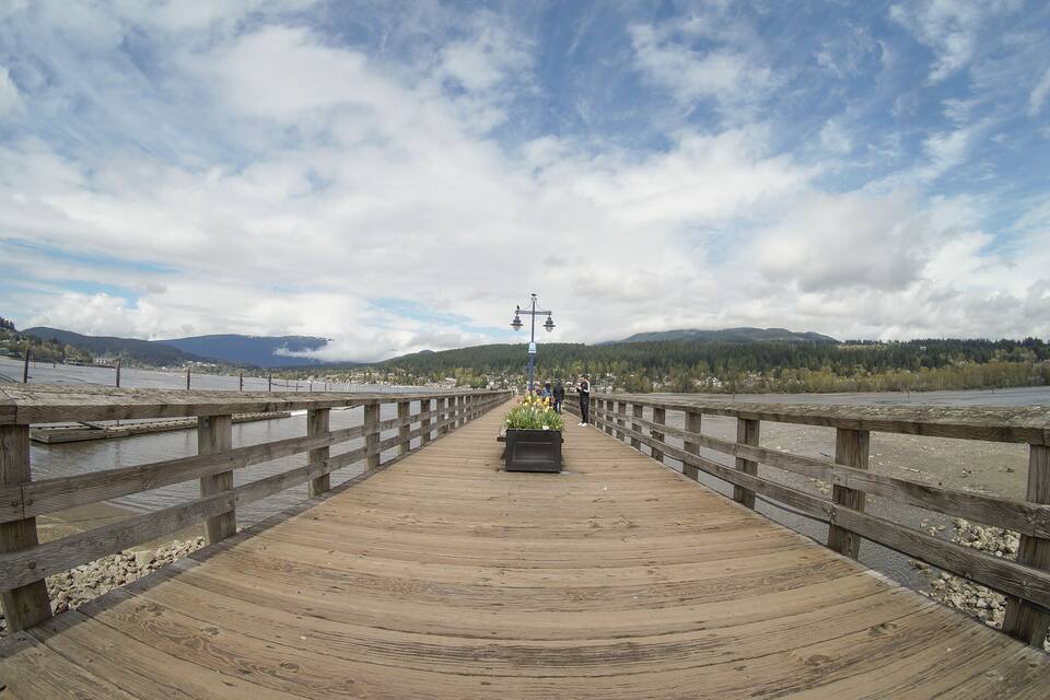 A wooden bridge over a river with a mountainous backdrop and a cloudy sky.