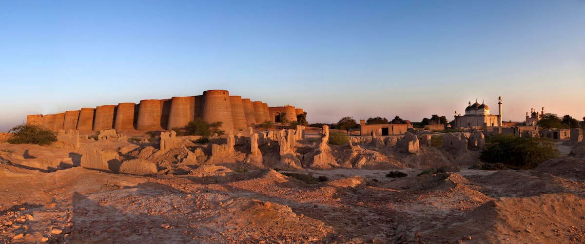 A historical fort with multiple towers, a mosque with a dome and minarets, and a desert landscape with sparse vegetation.