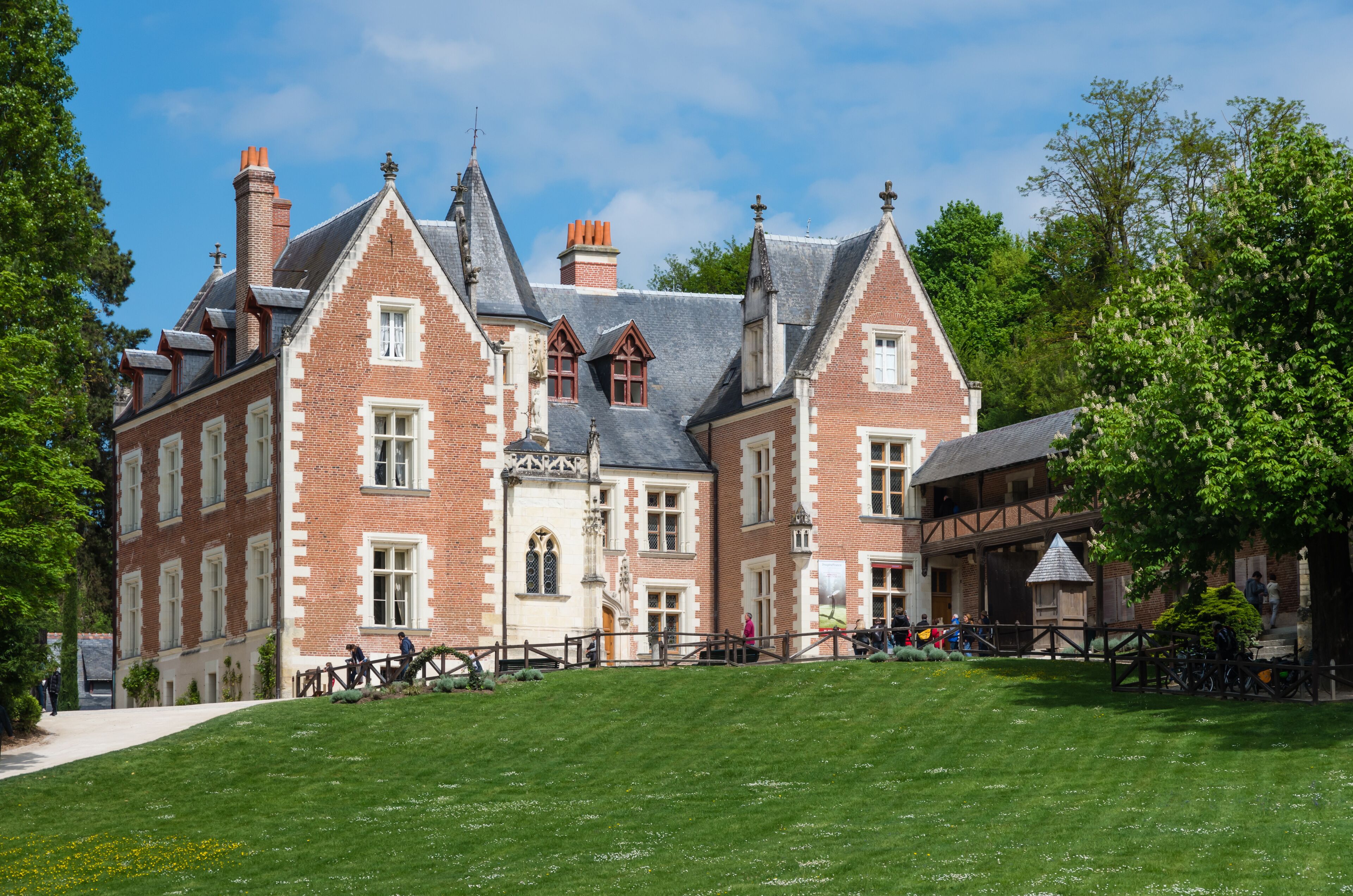 A large, historic brick building with multiple chimneys and a steeply pitched roof, surrounded by lush green lawns and trees.
