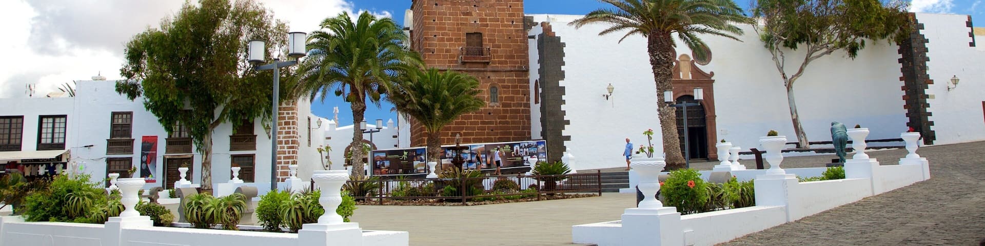 A historic clock tower with a white facade and a red brick base, surrounded by palm trees and white walls.