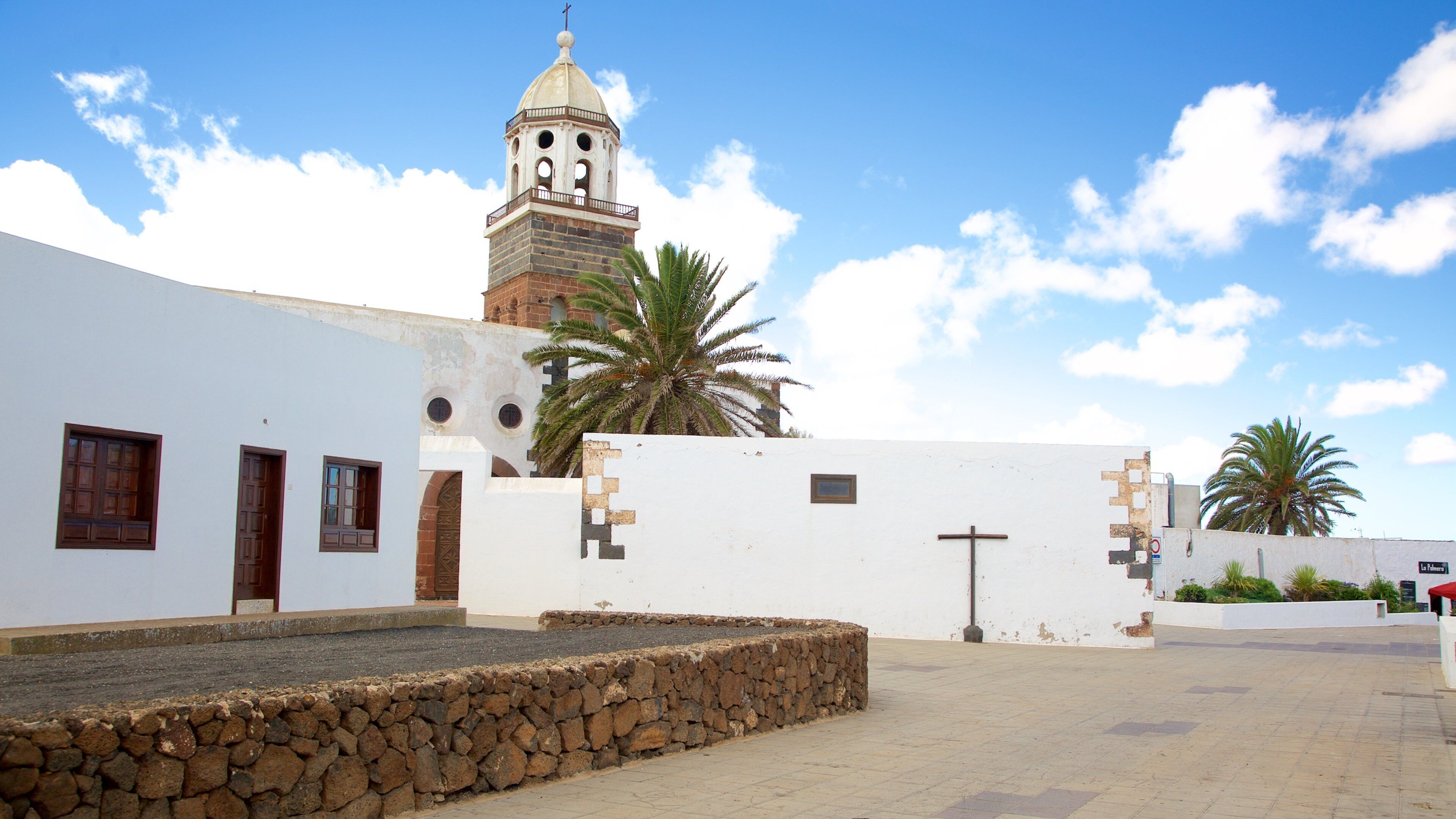 A white building with a stone wall, a palm tree, and a church tower.