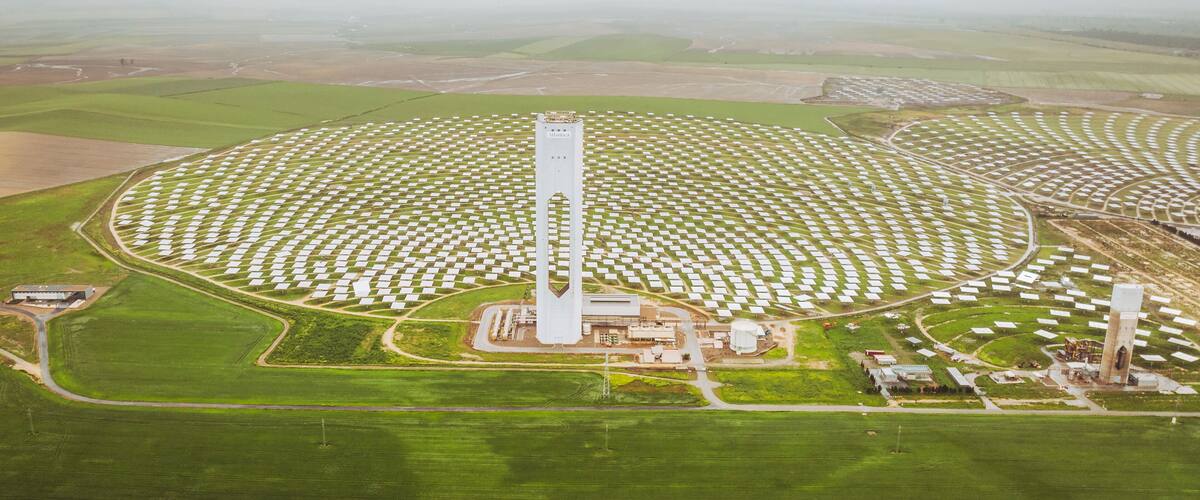 An aerial view of a solar farm with numerous solar panels arranged in a circular pattern, surrounded by green fields and a central tall tower.