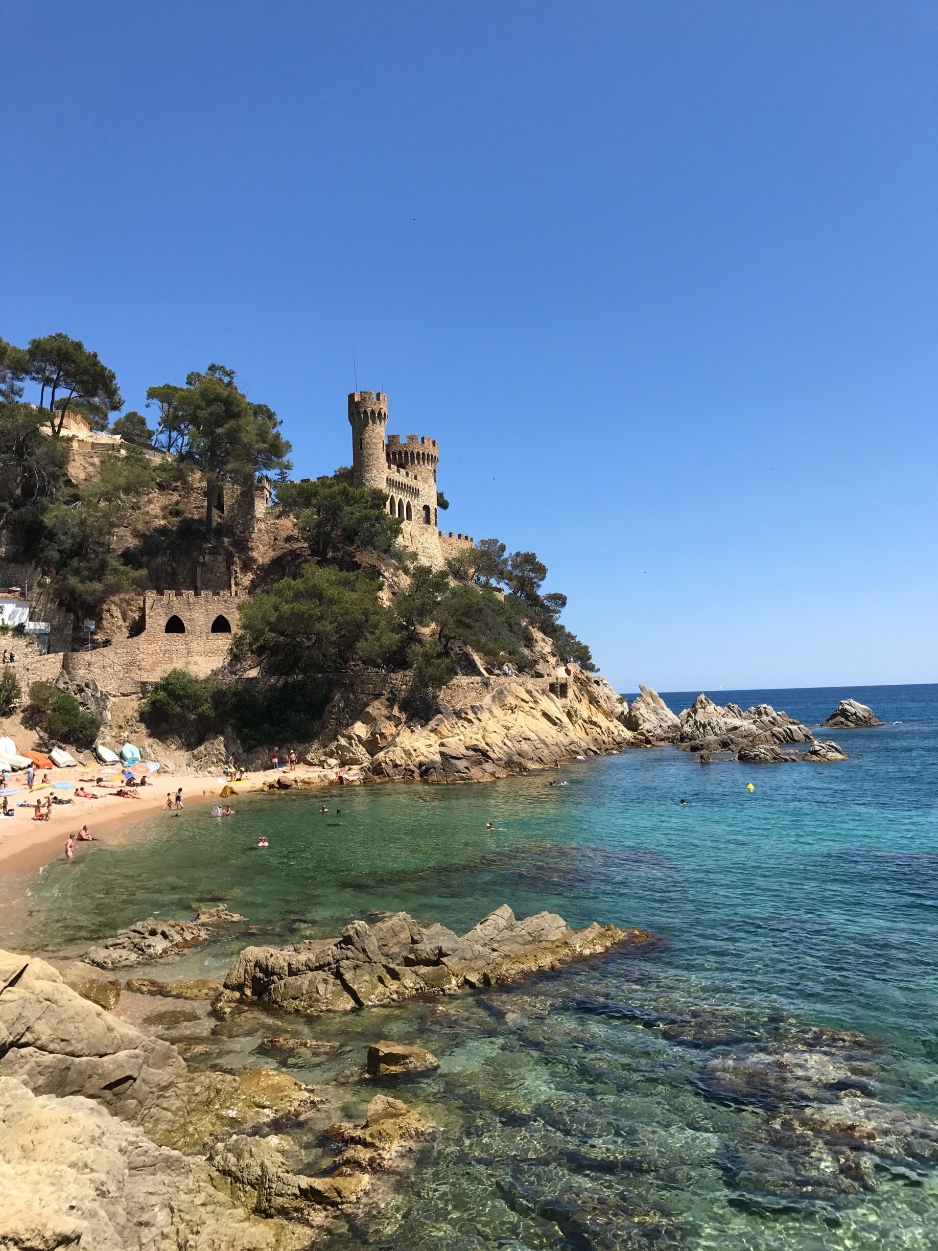 A coastal castle on a rocky cliff overlooking a clear blue sea with people swimming and sunbathing on the beach.