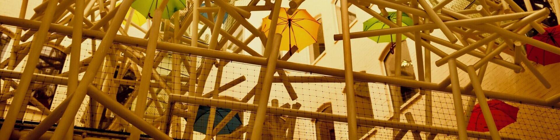 A complex structure of white metal beams with colorful umbrellas hanging from it.